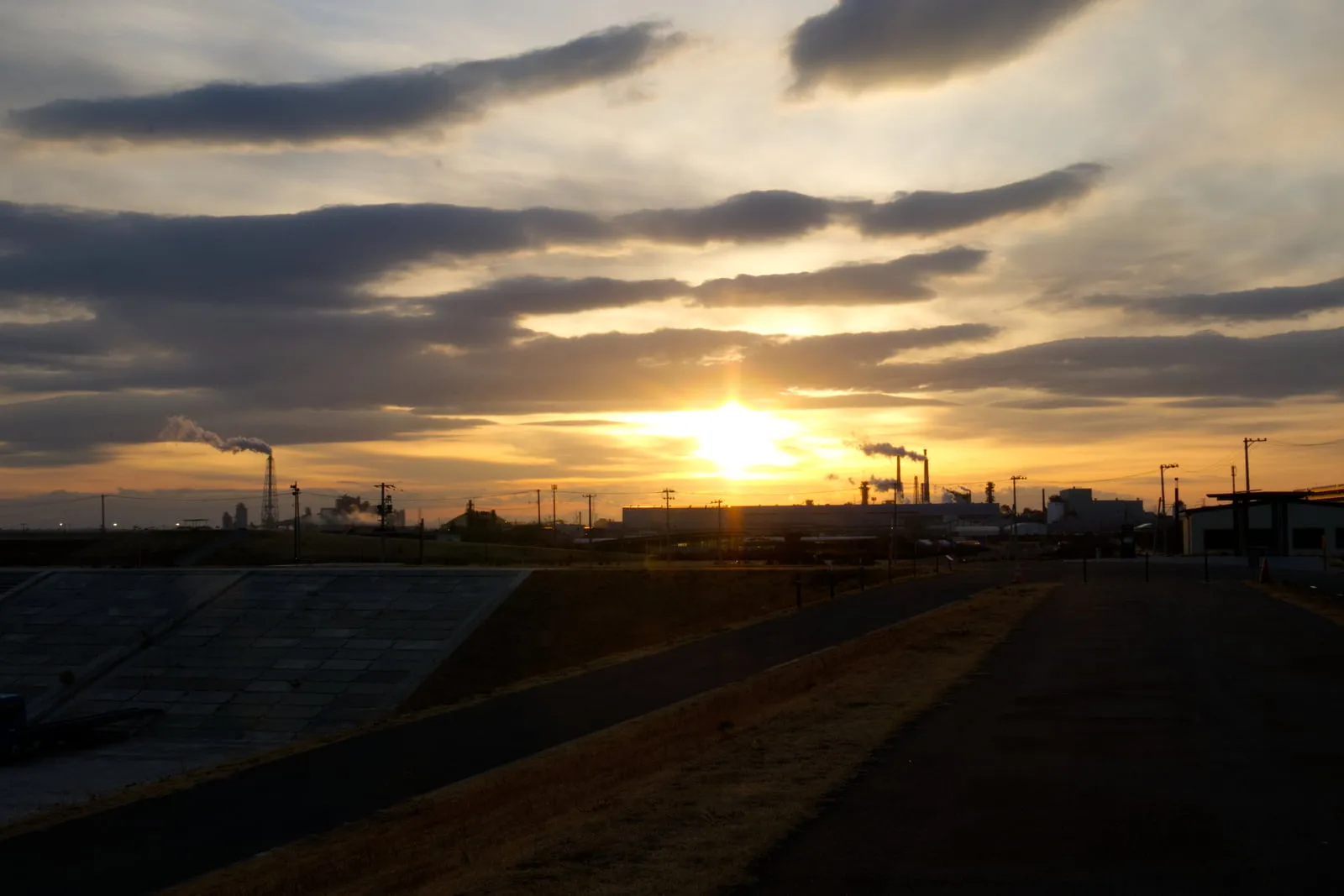 石巻市南浜マリーナそばの旧北上川土手から見る夕陽と、工場の煙突からのぼる煙の写真